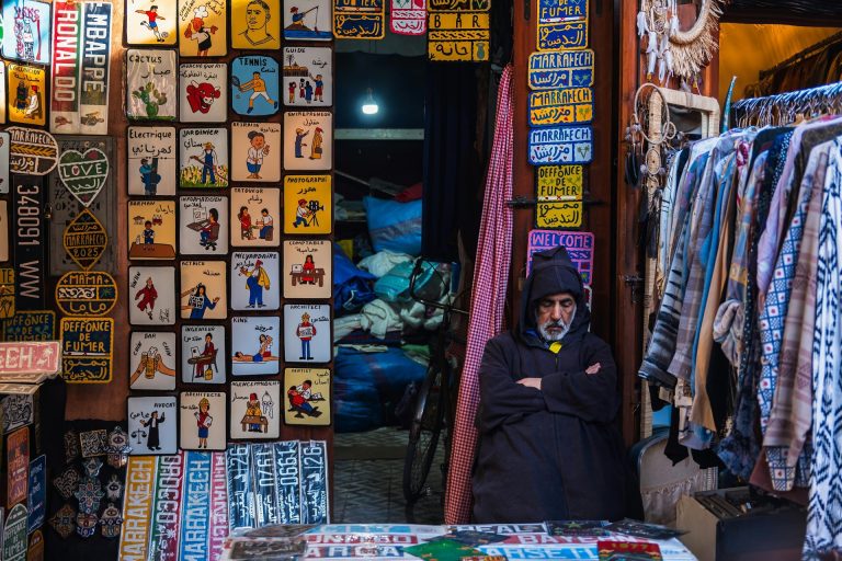 A vendor in Marrakech souk, surrounded by vibrant signs and garments, embodies local culture.