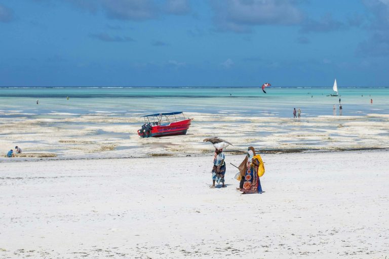 Women walking on a beach in Africa with a vibrant red boat and azure sea.