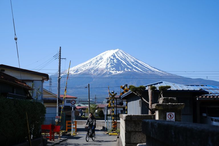 A person cycling near a railway crossing with Mount Fuji in the background under a clear blue sky.