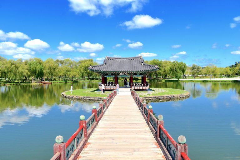 A serene pavilion on a pond's islet in a lush park under a clear blue sky.
