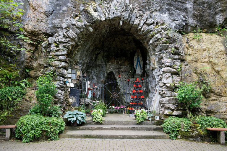 Rock grotto with Virgin Mary statue and vibrant red candles surrounded by lush greenery in Sarnen, Switzerland.