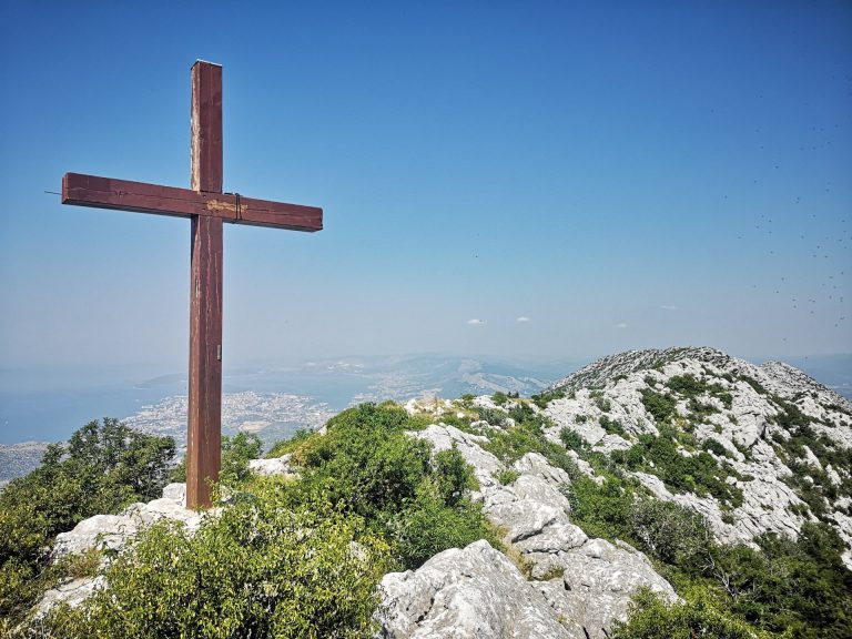 A scenic view of a wooden cross atop a rocky hill with coastal views, set under a blue sky.