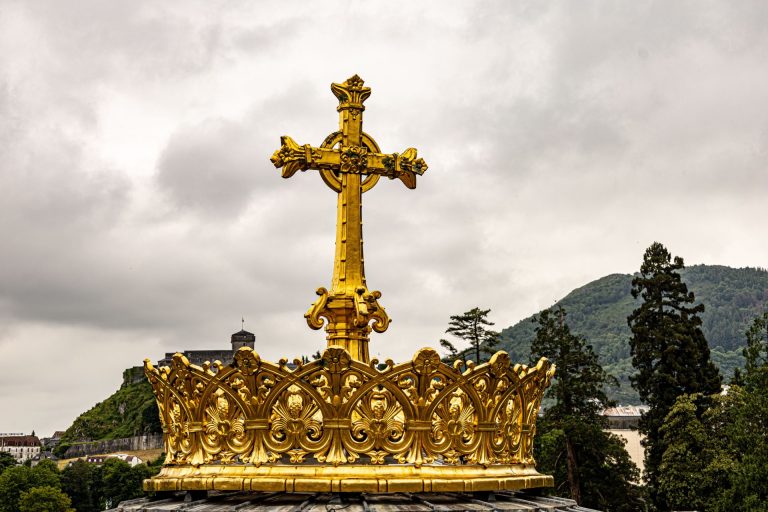 A majestic golden cross and crown sculpture stands against a cloudy sky with lush hills in the background.