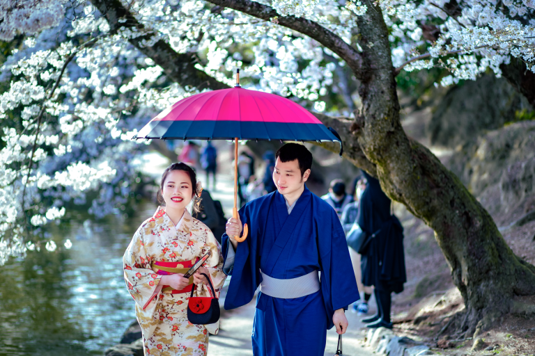 A couple in traditional kimonos walk under cherry blossoms holding an umbrella, symbolizing romance.