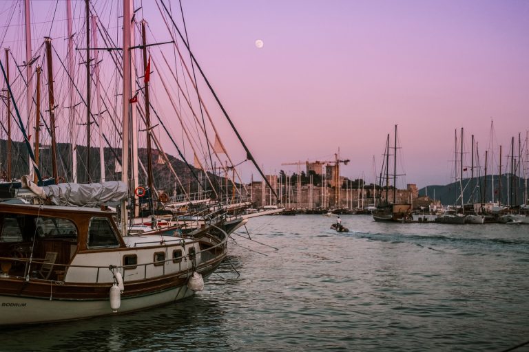 Scenic view of sailboats at dusk near a coastal castle with a serene pink sky.