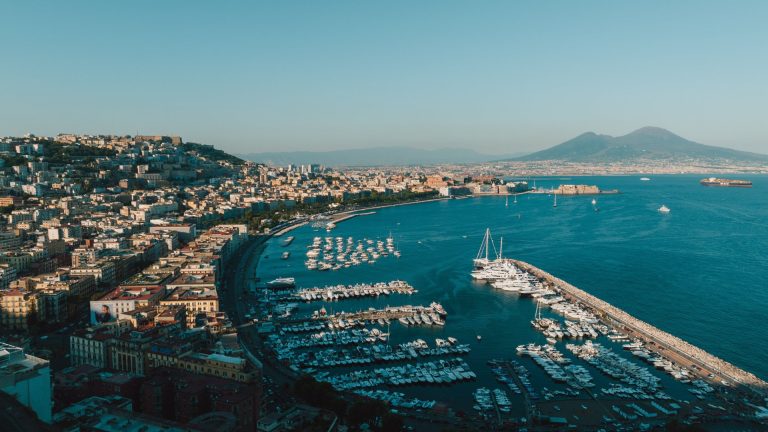 Aerial view of Naples harbor with boats and city skyline under clear blue sky.
